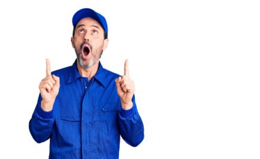 Tamirci üniforması giyen orta yaşlı yakışıklı adam şaşırmış ve şaşırmış bir şekilde yukarı bakıyor ve parmaklarıyla işaret ediyor ve kolunu kaldırıyor.. 