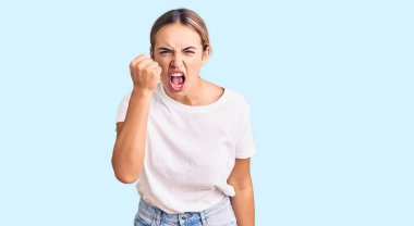 Genç, güzel, sarışın, rahat beyaz tişört giyen kızgın ve kızgın yumruk kaldıran sinirli ve öfkeyle bağıran bir kadın. öfke ve agresif konsept. 