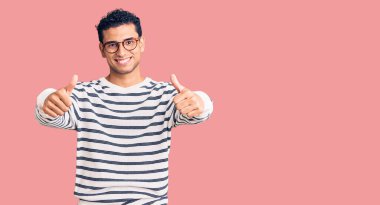 İspanyol asıllı yakışıklı genç adam gündelik giysiler giyiyor ve gözlük takıyor. Elle olumlu jest yapıyor, gülümsüyor ve başarı için seviniyor. kazanan jest. 