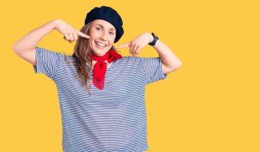 Fransız beresi takan, çizgili tişört giyen, gülümseyen, parmaklarını, dişlerini ve ağzını gösteren genç, güzel sarışın bir kadın. diş sağlığı kavramı. 