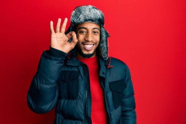 Sakallı, kışlık şapkalı, ceketli, gülümseyen Afro-Amerikalı genç adam el ve parmaklarıyla tamam işareti yapıyor. başarılı ifade. 