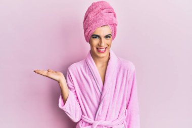 Kadın elbisesi giyen genç adam duş havlusu ve bornoz giyerek makyaj yapıyor neşeli bir şekilde gülümsüyor ve avuç içi kameraya bakıyor.. 