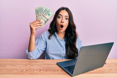 Ofiste çalışan güzel esmer genç bir kadın elinde dolarlarla korkmuş ve şaşırmış bir şekilde şaşkınlık içinde ağzı açık, inançsız bir yüz. 
