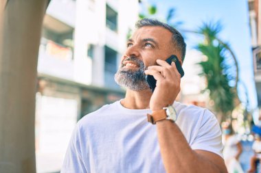 Orta yaşlı, kır saçlı, mutlu gülümseyen bir adam şehirde akıllı telefon kullanıyor.