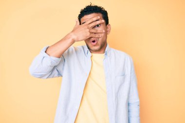 Genç, yakışıklı İspanyol adam sarı arka planda dikiliyor. Şoka girip yüzünü ve gözlerini eliyle kapatıyor. Korkmuş parmaklarından bakıyor. 