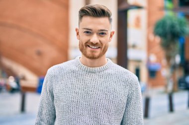 Genç beyaz adam gülümseyerek şehirde mutlu mesut duruyor..