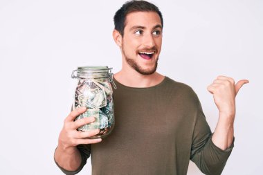 Genç yakışıklı adam elinde kavanozla başparmağıyla yan tarafa bakıyor mutlu bir şekilde ağzı açık gülümsüyor. 