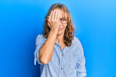 Orta yaşlı sarışın kadın günlük kıyafetler ve gözlük takıyor. Bir gözünü eliyle kapatıyor. Yüzünde kendine güvenen bir gülümseme ve sürpriz bir duygu var.. 