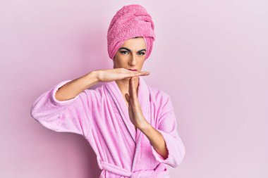 Kadın elbisesi giymiş genç bir adam kafasında duş havlusu ve bornozla el kol hareketi yapıyor, hayal kırıklığına uğramış ve ciddi bir yüz ifadesi var. 