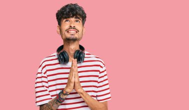 Kulaklıkla müzik dinleyen genç İspanyol adam elleri ile yalvarıyor ve dua ediyor. Yüzünde umut ifadesi var. Çok duygusal ve endişeli. Yalvarıyor. 