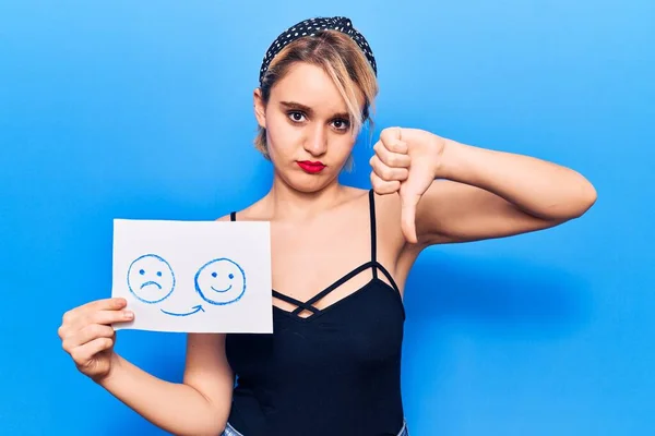 Young beautiful blonde woman holding sad to happy emotion paper with angry face, negative sign showing dislike with thumbs down, rejection concept