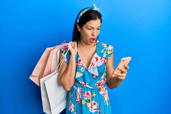 Jonge Latijnse Vrouw Houdt Boodschappentassen Smartphone Shock Gezicht Zoek Sceptisch — Stockfoto
