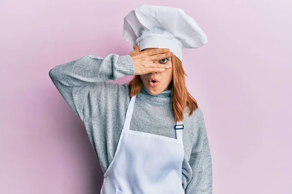 西班牙裔年轻女子身穿职业厨师制服 头戴安全帽 满脸通红 用手捂住眼睛 用手指看过去 很害怕 — 图库照片