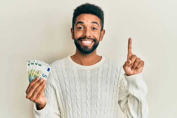 英俊而惊慌的男人留着胡子 手里拿着一堆5欧元的钞票 微笑着 带着一个想法或问题指手画脚 面带笑容 居首位 — 图库照片