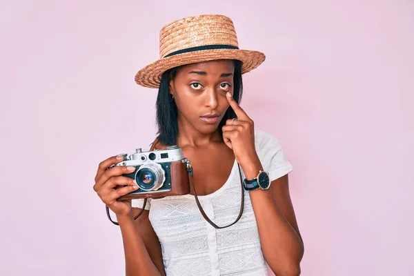 Young african american woman wearing summer hat holding vintage camera pointing to the eye watching you gesture, suspicious expression