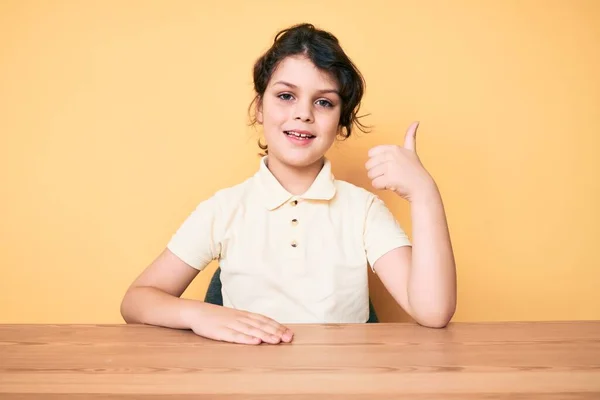 テーブルの上に座っているカジュアルな服を着てかわいいヒスパニック系の子供は幸せと肯定的な笑みを浮かべて 親指アップ優れた承認サインを行う — ストック写真