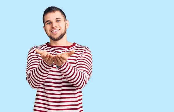 縞模様のセーターを着た若いハンサムな男が一緒に手の手のひらで笑顔を受信またはジェスチャーを与える 保持し保護する — ストック写真