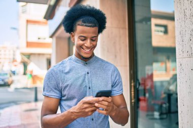 Genç Afrikalı Amerikalı adam şehirde akıllı telefon kullanarak mutlu bir şekilde gülümsüyor..