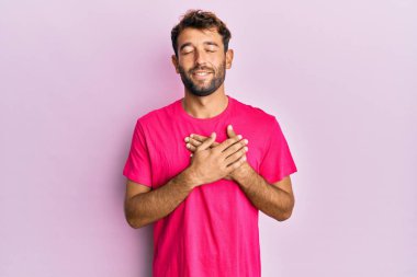 Pembe arka planda rahat pembe tişört giyen, elleri göğsünde, gözleri kapalı ve yüzünde minnet ifadesi olan yakışıklı bir adam. sağlık kavramı. 