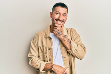 Dövmeleri olan, sıradan kahverengi kot ceket giyen, kameraya kendinden emin, kollarını kavuşturmuş ve çenesini kaldırmış gülümseyen yakışıklı bir adam. Olumlu düşünmek. 