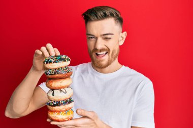 Genç kızıl saçlı adam elinde lezzetli renkli çöreklerle kameraya bakıyor. Seksi bir ifadeyle, neşeli ve mutlu bir yüzle.. 