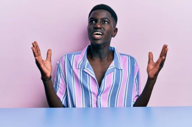 Gündelik giysiler giymiş genç Afro-Amerikan bir adam masaya oturmuş deli deli deli bağırıyor ve bağırıyor agresif bir ifade ve kollar havada. hüsran kavramı. 