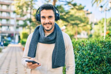 Parkta akıllı telefon ve kulaklık kullanarak mutlu gülümseyen genç İspanyol adam..