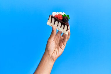 İspanyol adamın eli çikolatalı pastayı izole edilmiş mavi arka planda tutuyor..