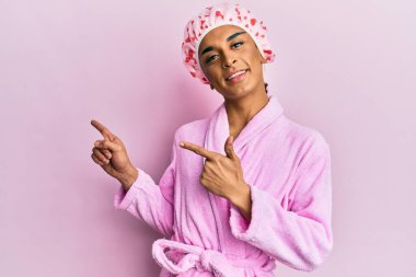 Duş havlusu şapkası ve bornoz takmış, gülümseyen ve kameraya bakan İspanyol bir adam iki eliyle ve parmaklarıyla kenarı işaret ediyor.. 
