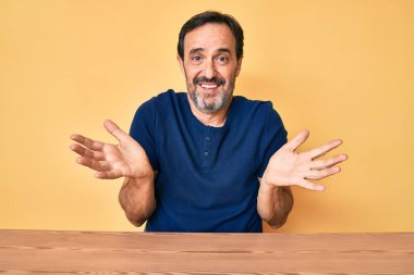 Orta yaşlı İspanyol bir adam, gündelik giysiler giyip masanın üzerinde elleri ve kolları havada, kafası karışmış bir şekilde oturuyor. şüphe kavramı. 