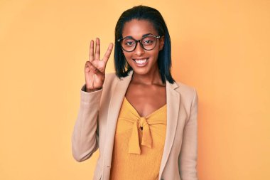 Genç afro-amerikan kadın iş kıyafetleri giyip üç numaralı parmaklarıyla işaret ederken kendinden emin ve mutlu bir şekilde gülümsüyor.. 