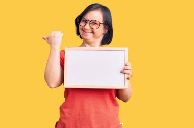 Down sendromlu esmer kadın elinde boş beyaz karatahta ile baş parmağını yana uzatıyor ve ağzı açık gülümsüyor. 