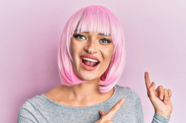 Pembe peruklu genç beyaz kadın gülümsüyor ve kameraya bakıyor. İki eli ve parmağı yana bakıyor.. 