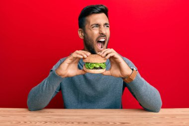 Yakışıklı İspanyol bir adam lezzetli klasik bir burger yiyor kızgın ve öfkeli çığlıklar atıyor öfkeyle bağırıyor. öfke ve agresif konsept. 