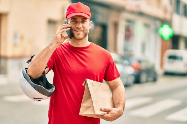 Beyaz atlı genç teslimatçı akıllı telefondan konuşuyor ve şehirde kağıt torba taşıyor..