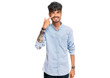 Latin asıllı genç bir adam, gündelik giysiler giyip el ve burnu işaret ediyor, neşeli bir şekilde gülümsüyor. güzellik konsepti 