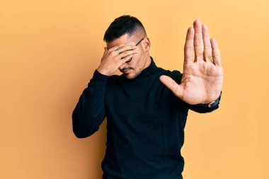 Balıkçı yaka kazak giyen, gözlük takan, gözlerini elleriyle kapayan ve üzgün ve korku ifadeli hareketler yapan yakışıklı bir adam. utangaç ve olumsuz kavram. 