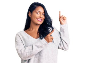 Uzun saçlı İspanyol kadın günlük kıyafetler giyiyor gülümsüyor ve kameraya bakıyor. İki eliyle ve parmaklarıyla kenarı işaret ediyor.. 