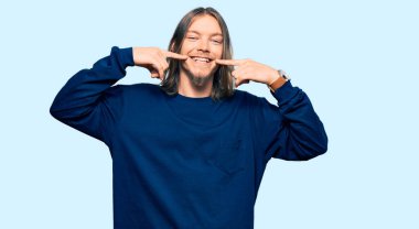 Uzun saçlı, sıradan bir süveter giyen, neşeli gülümseyen, parmaklarını, dişlerini ve ağzını gösteren yakışıklı beyaz bir adam. diş sağlığı kavramı. 