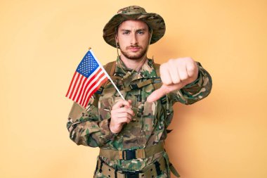 Kafkasyalı genç adam kamuflaj üniforması giyiyor. Elinde kızgın yüzlü Amerikan bayrağı var. Negatif işaretler baş parmakları aşağıda, reddedilme konsepti reddedildi. 