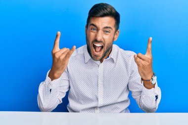 İş kıyafetleri giymiş yakışıklı İspanyol adam masaya oturmuş deli gibi bağırıyor elleri havada rock sembolü yapıyor. Müzik yıldızı. ağır müzik konsepti. 