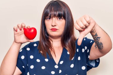 Kızgın yüzlü, kalbi tutan genç ve iri bir kadın. Negatif işaretler, baş parmakları aşağıda, reddedilme konsepti. 