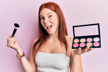 Genç kızıl saçlı kız elinde makyaj fırçası ve allık paleti ile çılgınlığı kutluyor ve şaşkınlık içinde gözleri açık heyecanla bağırıyor.. 
