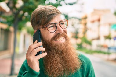 Kızıl saçlı genç İrlandalı adam mutlu bir şekilde gülümsüyor ve şehirdeki akıllı telefondan konuşuyor..