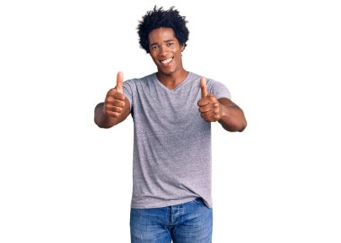Afro saçlı, sıradan kıyafetler giyen, el hareketleriyle olumlu jest yapan, gülümseyen ve başarı için mutlu olan yakışıklı Afro-Amerikan erkeği. kazanan jest. 