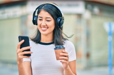 Genç İspanyol kadın kahve içiyor ve şehirde akıllı telefon ve kulaklık kullanıyor..