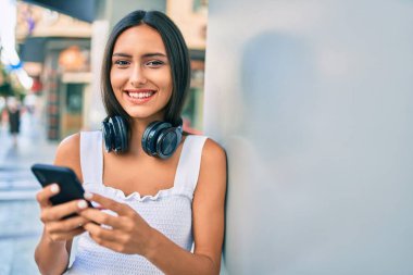 Duvara dayanmış akıllı telefon kullanan, kulaklık takan genç Latin kız..