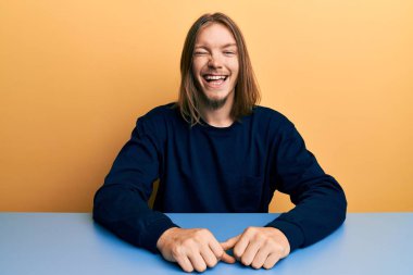 Uzun saçlı, yakışıklı beyaz adam. Masada oturmuş kameraya seksi bir ifadeyle bakıyor. Neşeli ve mutlu bir yüzü var.. 