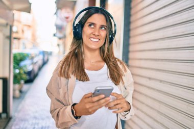 Beyaz kadın şehirde akıllı telefon ve kulaklık kullanarak mutlu bir şekilde gülümsüyor..