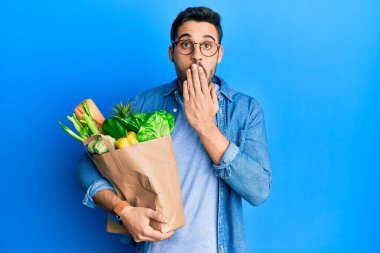 Elinde ekmek ve yiyecek dolu bir poşet tutan genç bir İspanyol, ağzı elle kapatılmış, şok olmuş ve hata yapmaktan korkan. şaşırmış ifade 